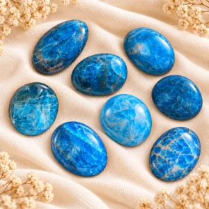 Ensemble de 8 galets d’apatite bleue naturelle de Madagascar, présentés sur un tissu beige doux avec de petites fleurs séchées.